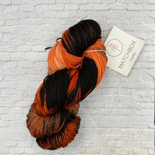 Load image into Gallery viewer, Braided skein of orange and black yarn with a Matchbox Yarn label on a white brick background