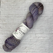 Load image into Gallery viewer, Gray and purple skein of yarn with a label on a light gray brick background