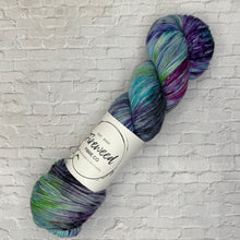 Load image into Gallery viewer, Multicolored skein of yarn with a label on a light gray brick background

