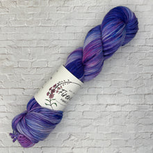 Load image into Gallery viewer, Purple skein of yarn with a label on a white brick background
