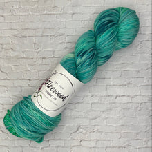 Load image into Gallery viewer, Green skein of yarn with a label on a white brick background
