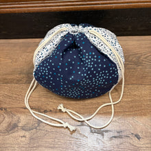 Load image into Gallery viewer, A small fabric drawstring pouch with a navy blue body covered in small white and light blue star patterns, contrasted with white and navy polka dot fabric panels and cream-colored drawstring cords, resting on a wooden surface.
