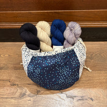 Load image into Gallery viewer, A fabric drawstring bag filled with four skeins of yarn in black, cream, navy, and lavender, placed on a wooden floor in front of a wooden wall.
