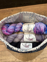 Load image into Gallery viewer, Three skeins of Fingertone yarn in different colors (purple, pink, and gray) arranged inside a fabric storage basket on a wooden surface.
