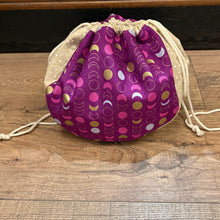 Load image into Gallery viewer, A fabric drawstring pouch with a purple front panel featuring a gold, pink, and white moon phase pattern, and a beige dotted reverse side, resting on a wooden floor.
