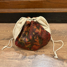 Load image into Gallery viewer, A small drawstring pouch with a cream-colored fabric body and a dark red, brown, and green patterned front panel, resting on a wooden surface.
