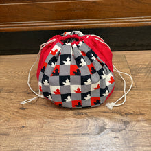 Load image into Gallery viewer, A small drawstring storage bag with a red, gray, and black checkered pattern featuring red and white maple leaves, resting on a wooden floor.
