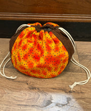 Load image into Gallery viewer, A fabric drawstring pouch bag with a brown side panel and a front panel featuring a yellow, orange, and red spotted pattern, resting on a wooden floor.
