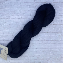 Load image into Gallery viewer, Dark blue skein of yarn with a label on a light brick wall background
