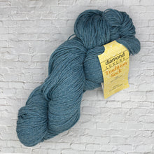 Load image into Gallery viewer, Blue skein of yarn with a yellow label on a light gray brick wall background

