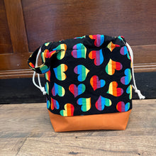 Load image into Gallery viewer, Drawstring bag with rainbow heart pattern on black fabric and orange base, placed on a wooden floor.
